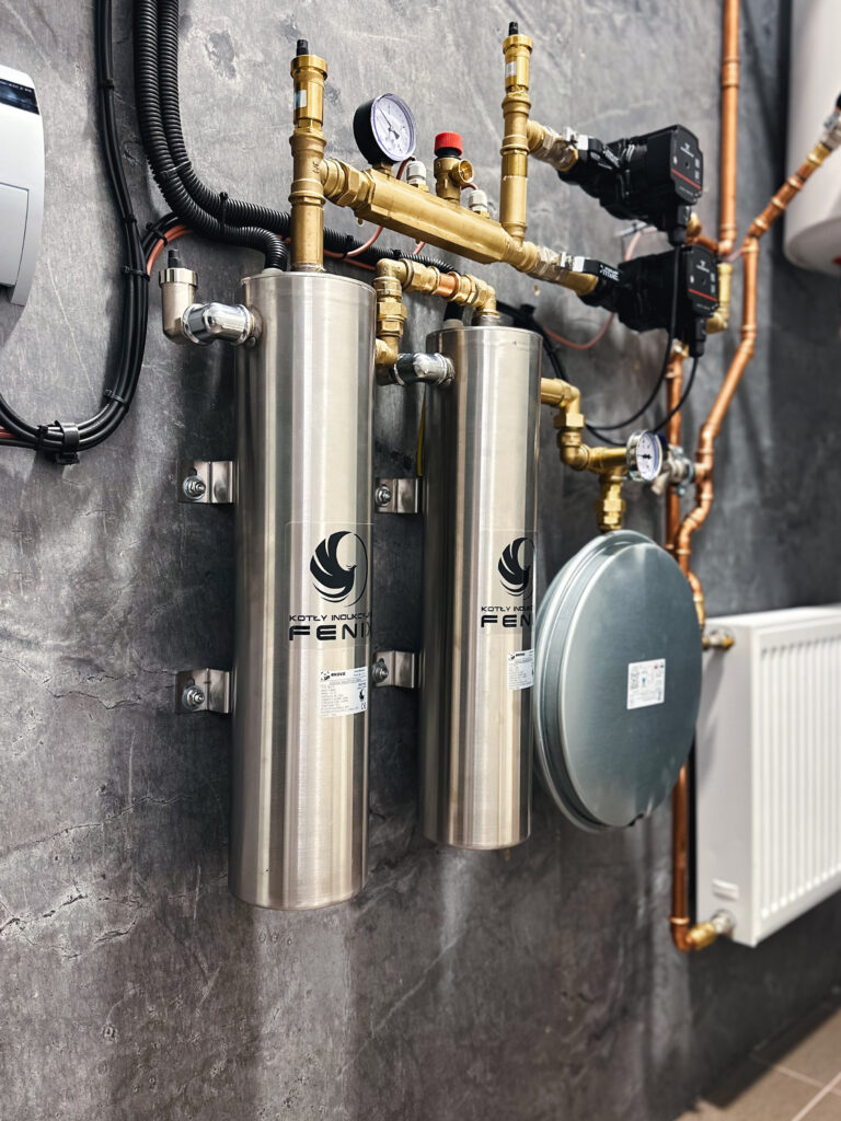 Instalacja kotłów indukcyjnych Fenix o mocy 9 kW. Montaż został wykonany na szarej ścianie. Obok widać grzejnik koloru białego oraz boiler.