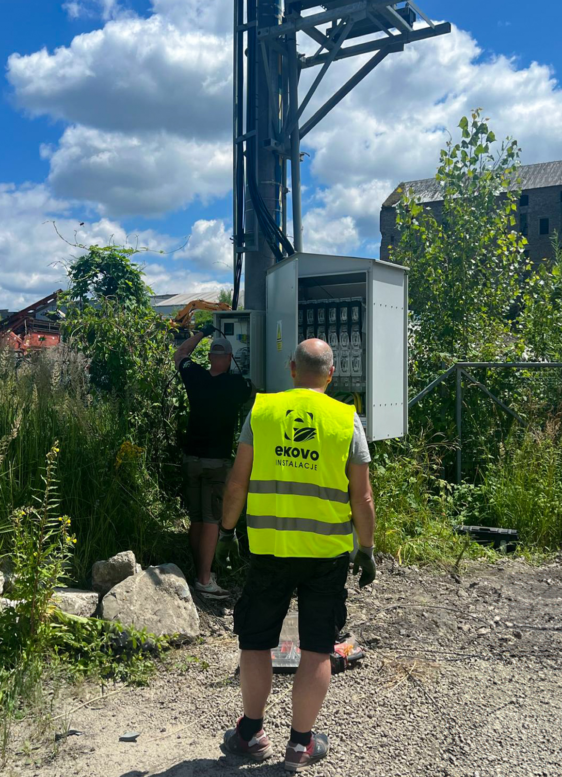 Pracownicy Ekovo w trakcie robót elektrycznych. Pierwszy mężczyzna po lewej trzyma kabel. stoi tyłem. Jest ubrany na czarno i ma szarą czapkę z daszkiem. Drugi mężczyzna również odwrócony tyłem.Jest ubrany w czarne szorty szarą koszulkę. Ma również kamizelkę z napisem Ekovo Instalacje. W tle zachmurzone niebo oraz krzewy.